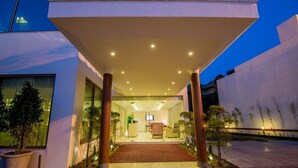 A modern building entrance with a covered walkway, potted plants, and a reception area visible inside.