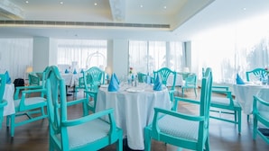 A dining area with turquoise chairs and white tablecloths, large windows, and a chandelier.