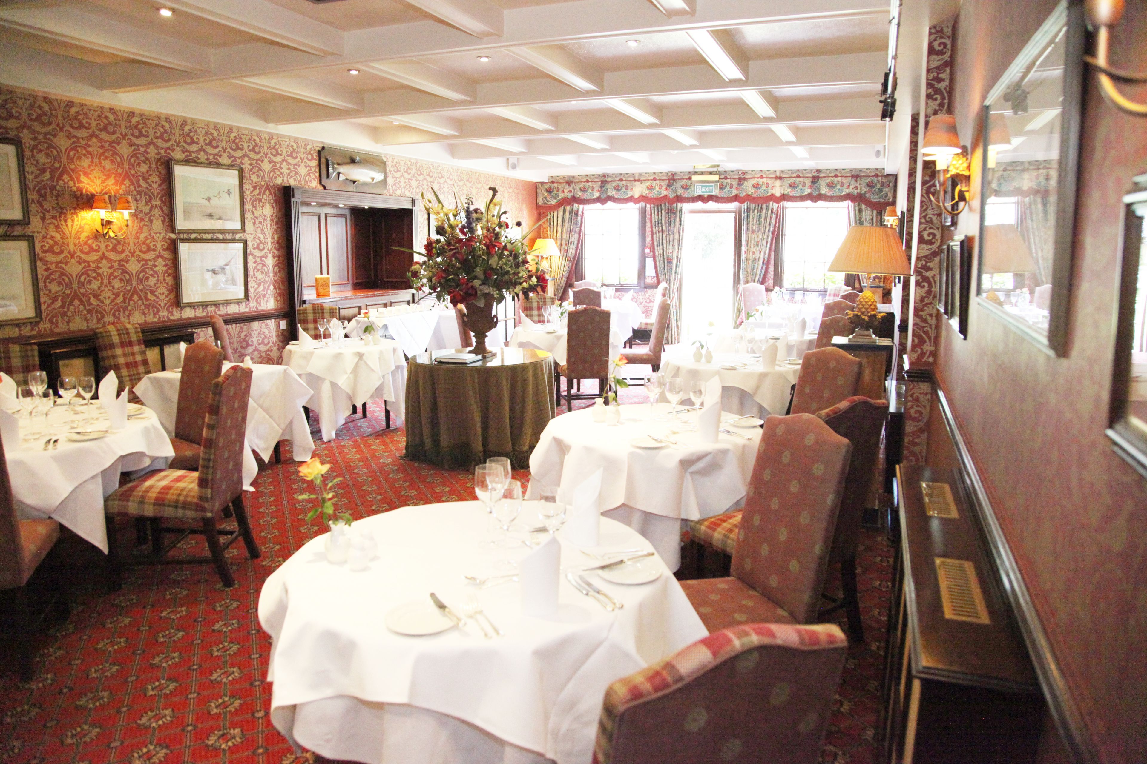 A dining area with round tables set for four, each with white tablecloths and silverware. There are floral centerpieces on the tables, and the room has patterned wallpaper and a red carpet.