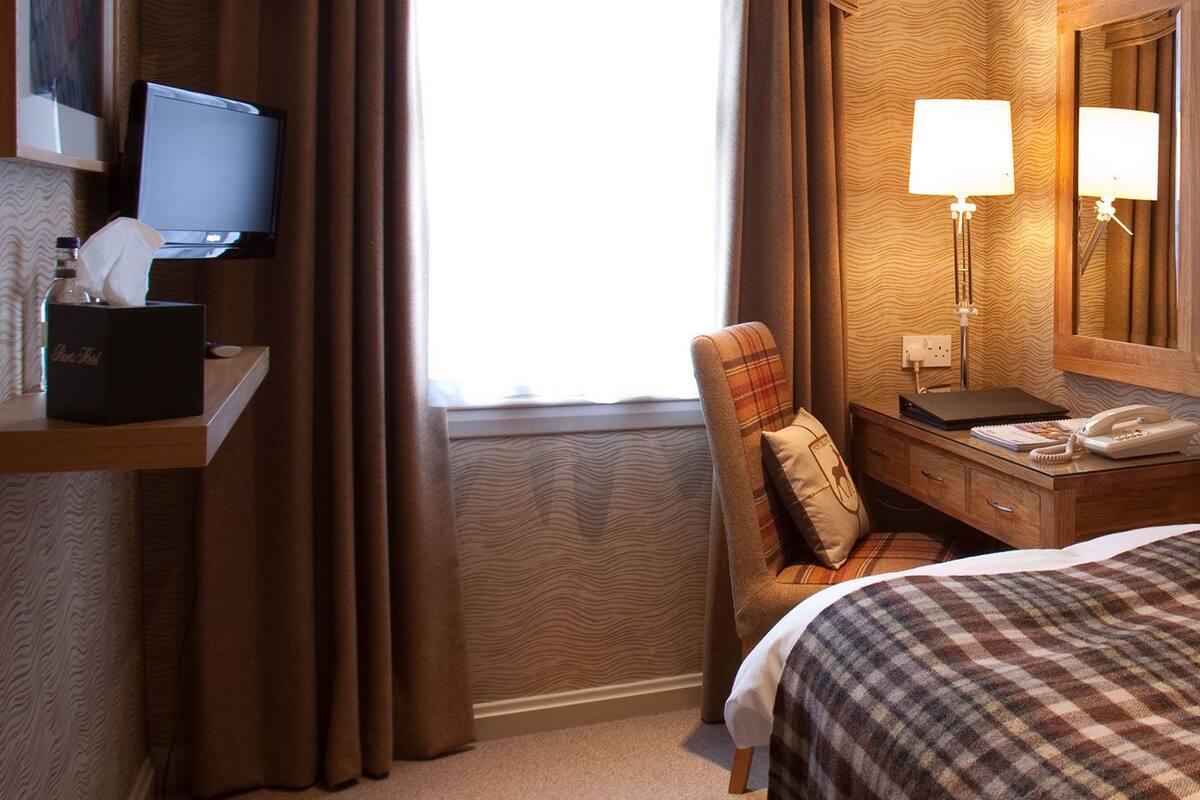 A hotel room with a bed, a desk with a lamp, a telephone, and a television on a stand.