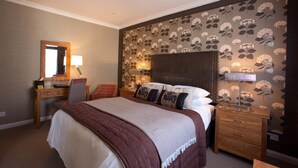 Habitación de hotel con cama, escritorio, silla, mesita de noche y papel tapiz floral.