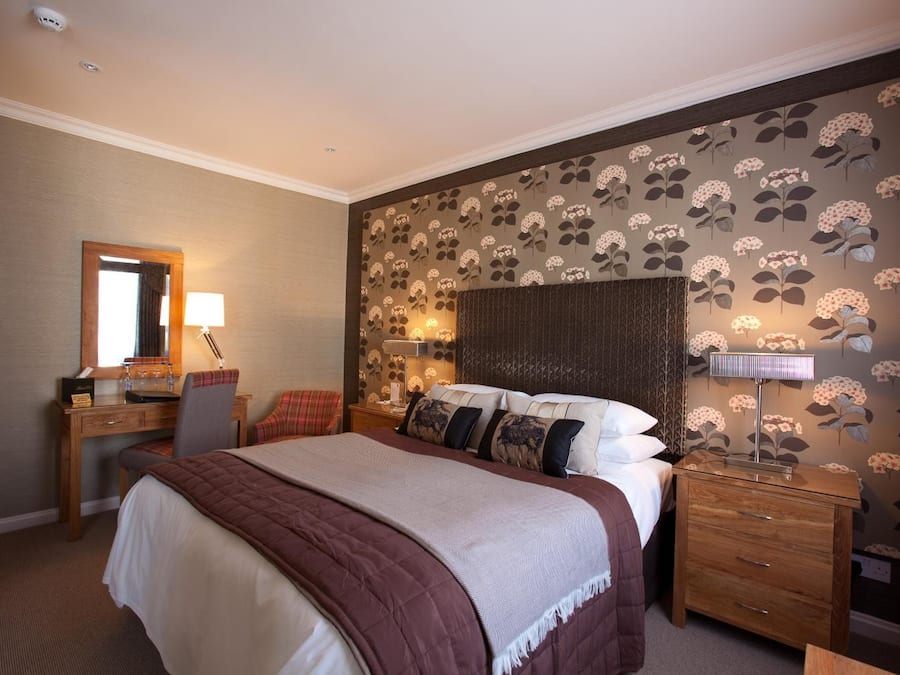 A hotel room with a bed, a desk, a chair, a nightstand, and a wall with floral wallpaper.
