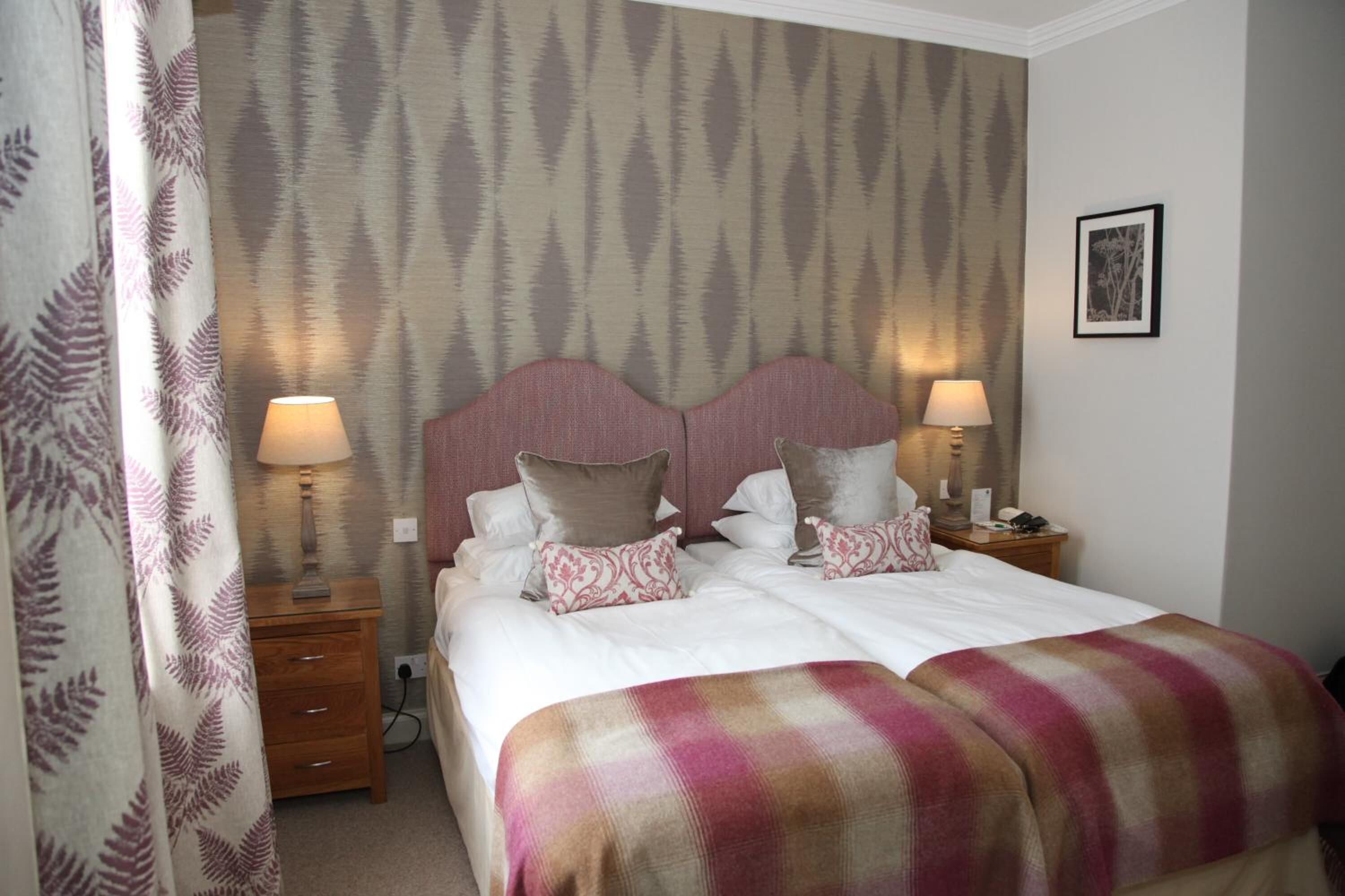 A double bed with a pink and beige blanket, two bedside tables with lamps, a nightstand, and a framed picture on the wall.