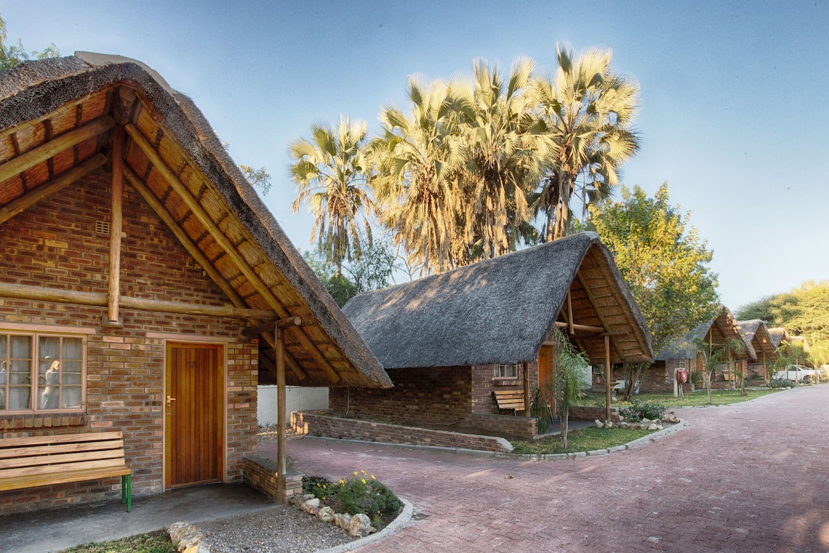 A rustic lodge with thatched roofs and brick walls, featuring a paved driveway and palm trees in the background.