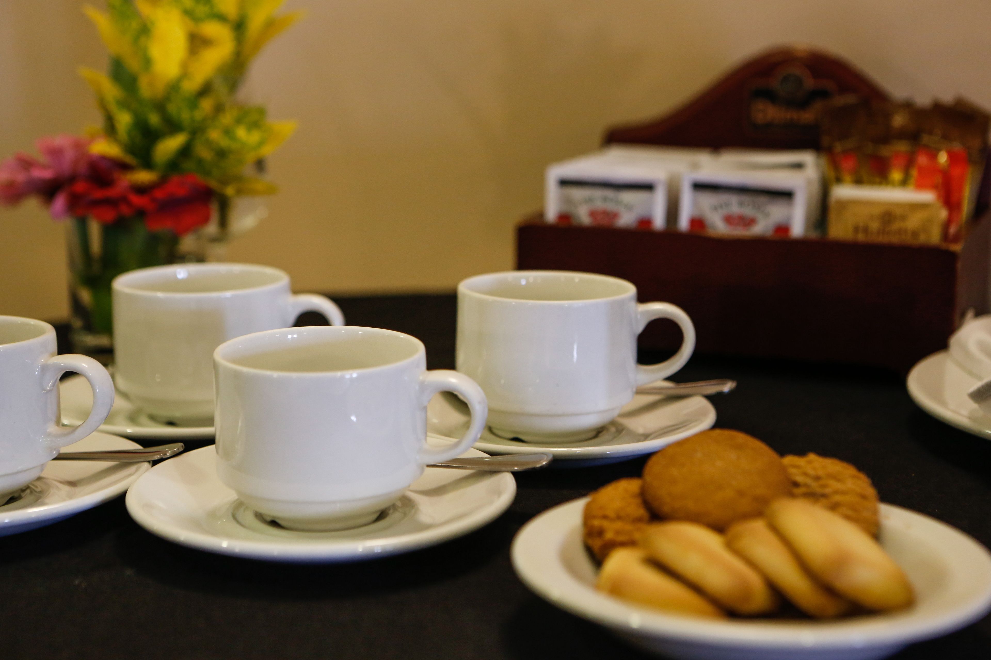 A table with white cups and saucers, a plate with cookies, and a box of tea bags in the background.