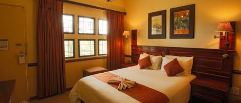 Une chambre d’hôtel avec un grand lit, une tête de lit en bois, un lustre, une fenêtre avec des rideaux et deux tableaux encadrés accrochés au mur.