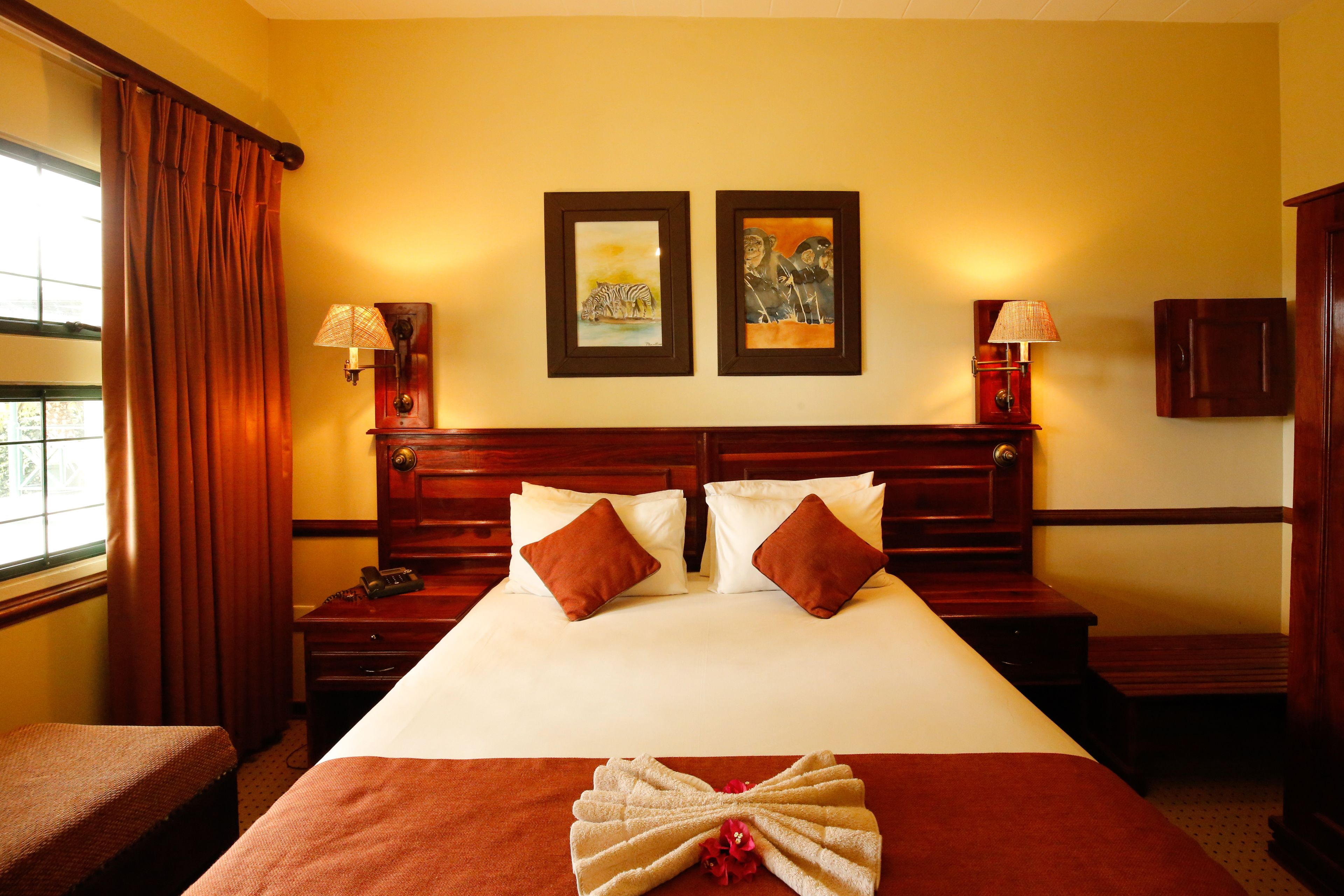 A hotel room with a large bed, two nightstands, a wooden headboard, two framed pictures on the wall, and a window with curtains.