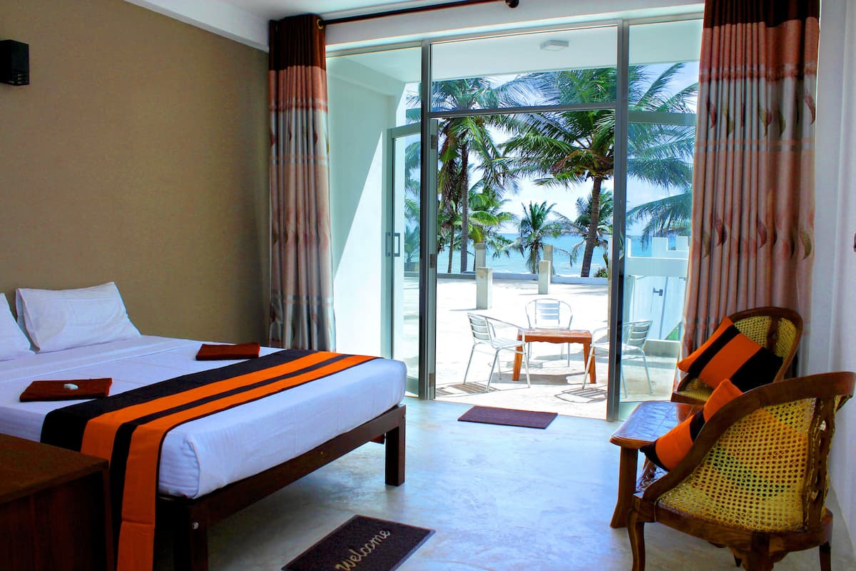 Een hotelkamer met een bed, een houten stoel en uitzicht op het strand en palmbomen door een glazen deur.
