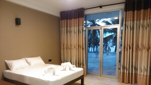 Habitación de hotel con cama, mesita de noche, ventana con cortinas y vistas a palmeras y una playa.