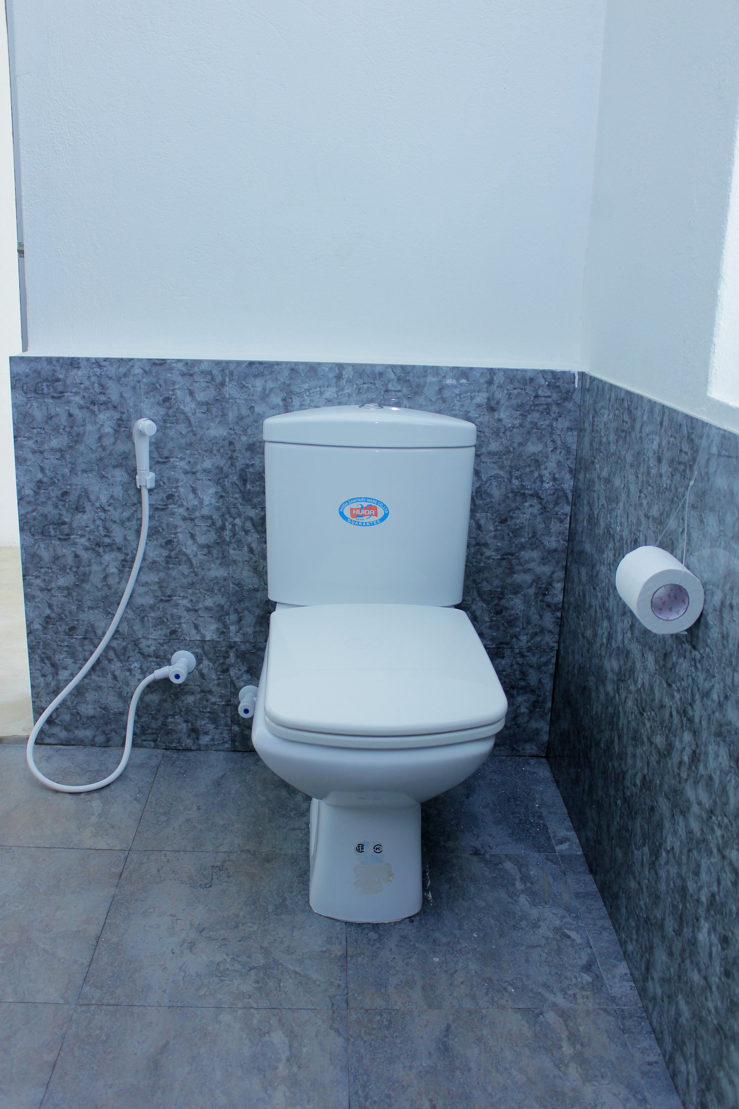 Un inodoro blanco con un adhesivo azul, un dispensador de papel higiénico y una manguera.
