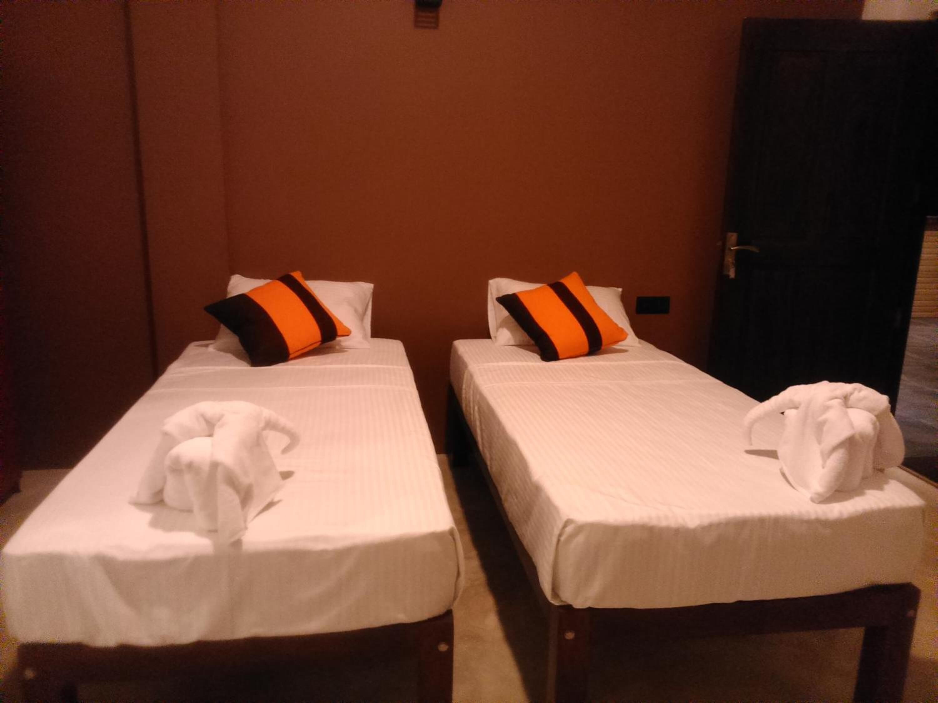Two single beds with white linens and orange and black striped pillows, a white towel on each bed, and a wooden door in the background.