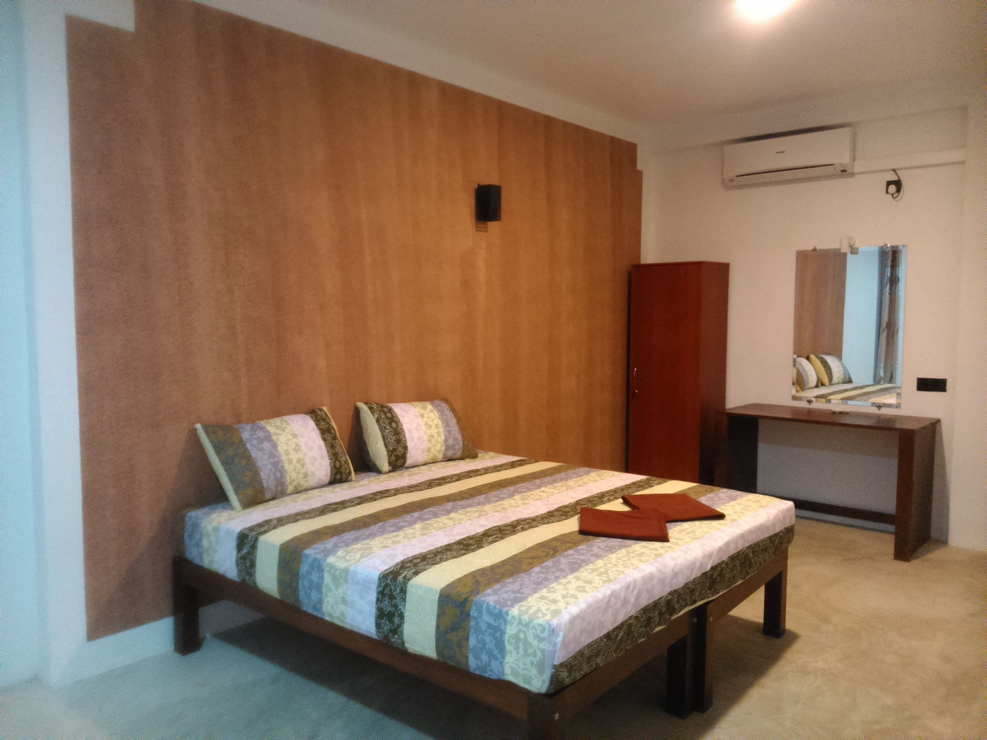 A bedroom with a bed, a wooden wardrobe, a desk, and a mirror.