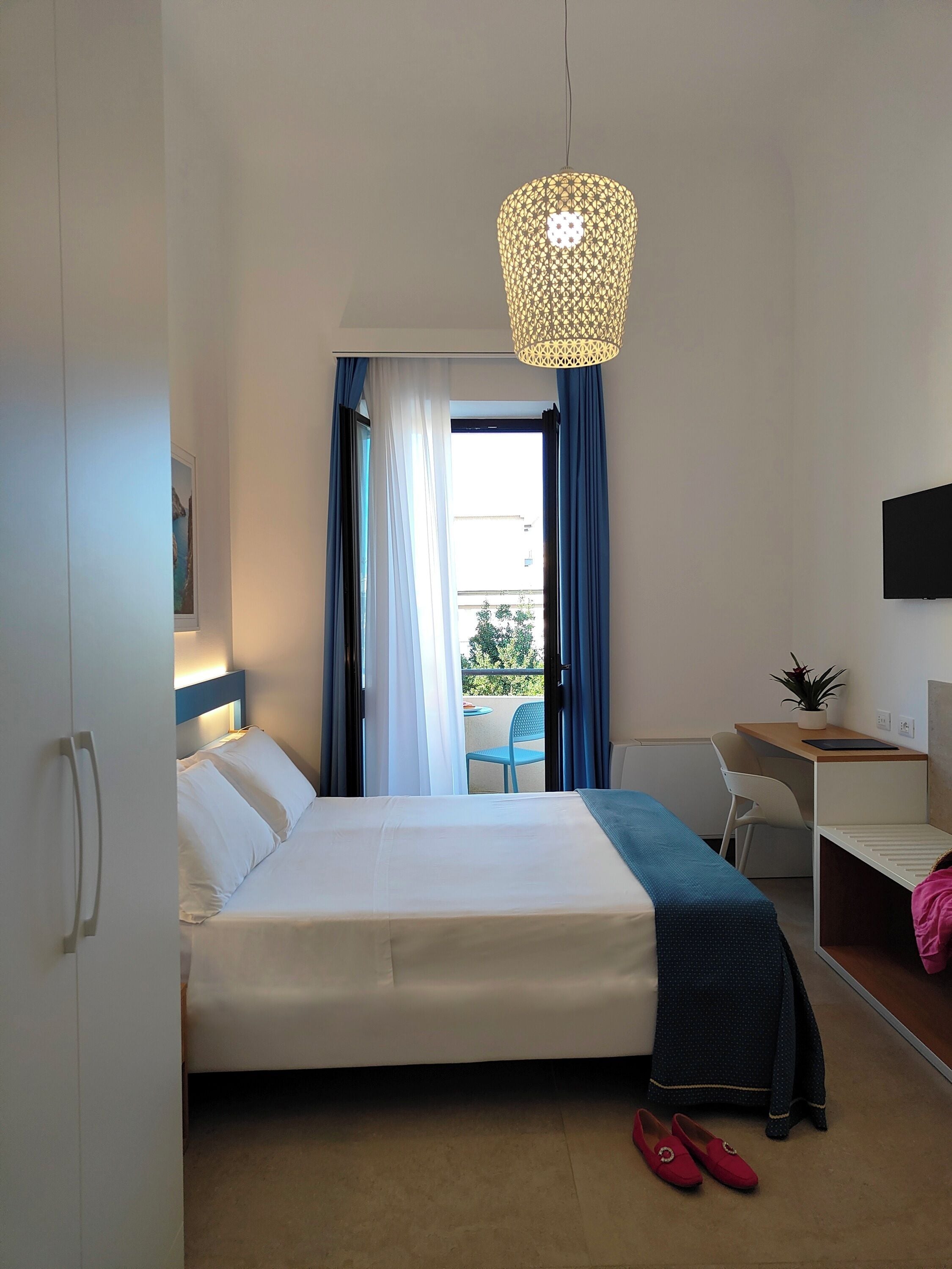A hotel room with a bed, a desk, a chair, a TV, and a balcony with blue curtains.