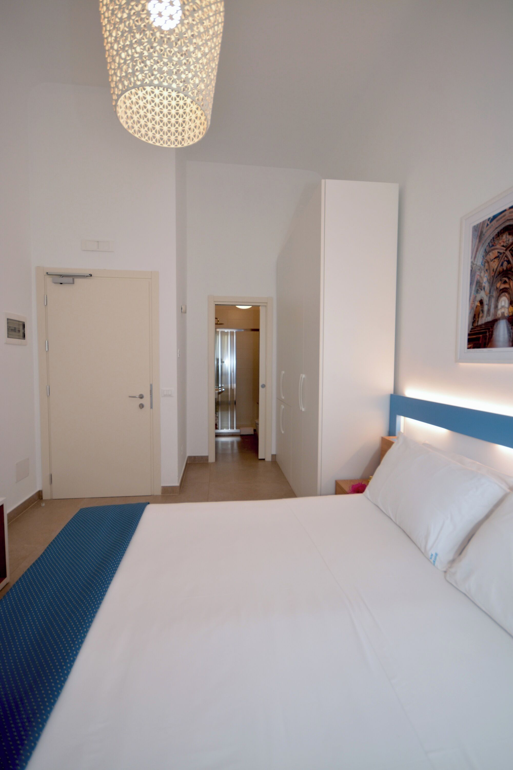 A hotel room with a bed, a door, a hallway, and a light fixture.