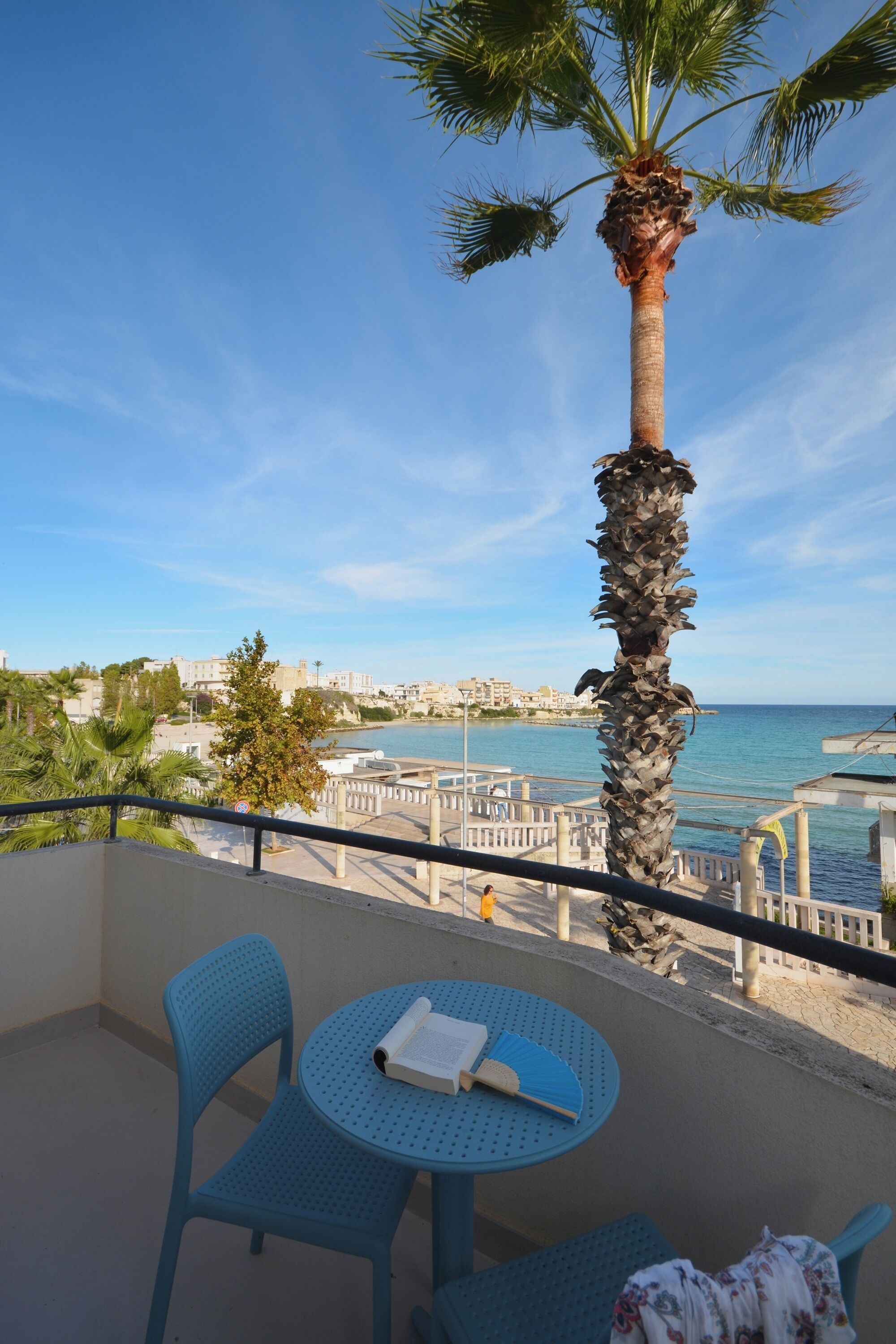 A balcony with a view of the sea, a palm tree, and a table with a book and a fan.
