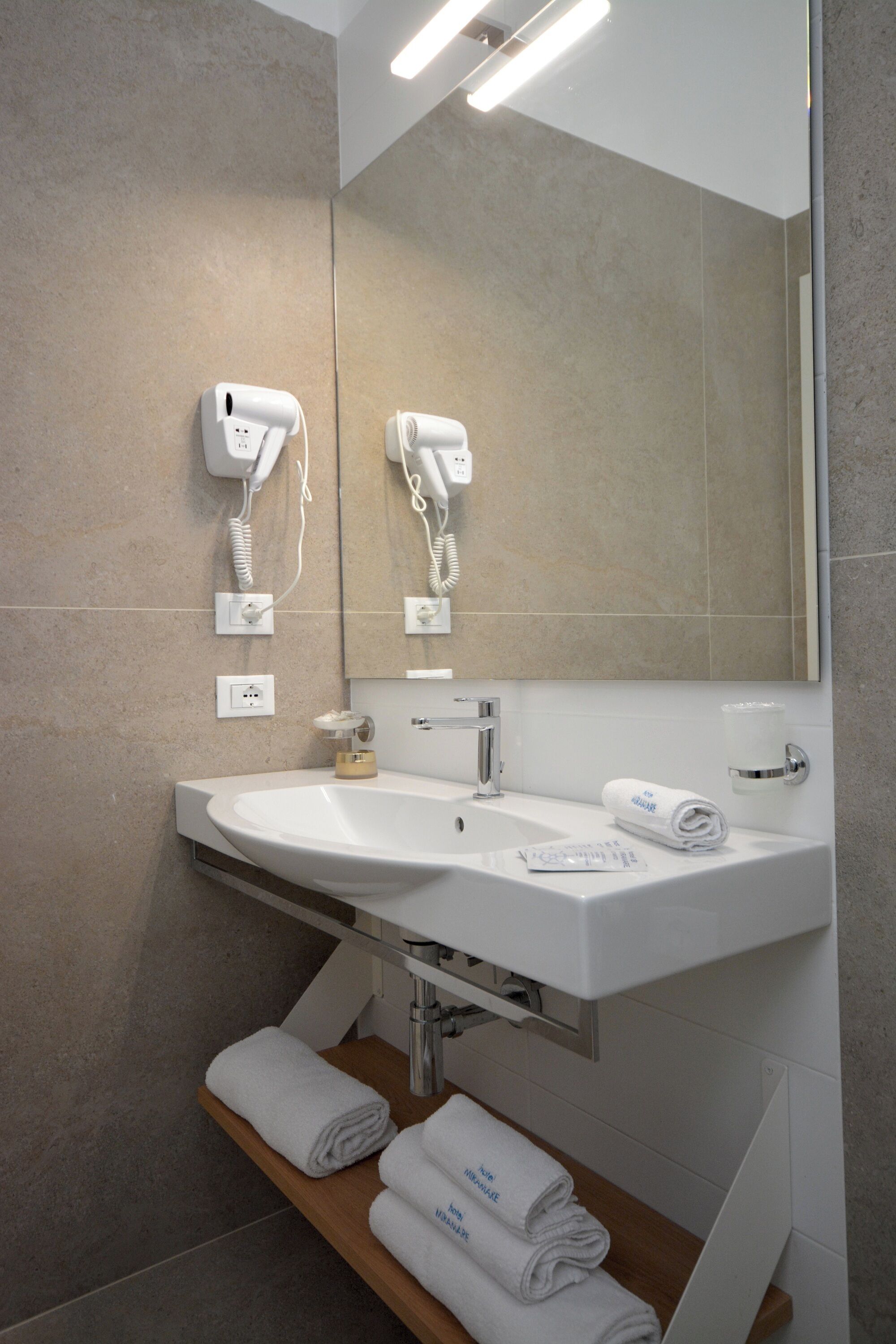 Un baño moderno con un lavamanos grande, dos secadores de pelo montados en la pared, un espejo y un estante de madera con toallas.
