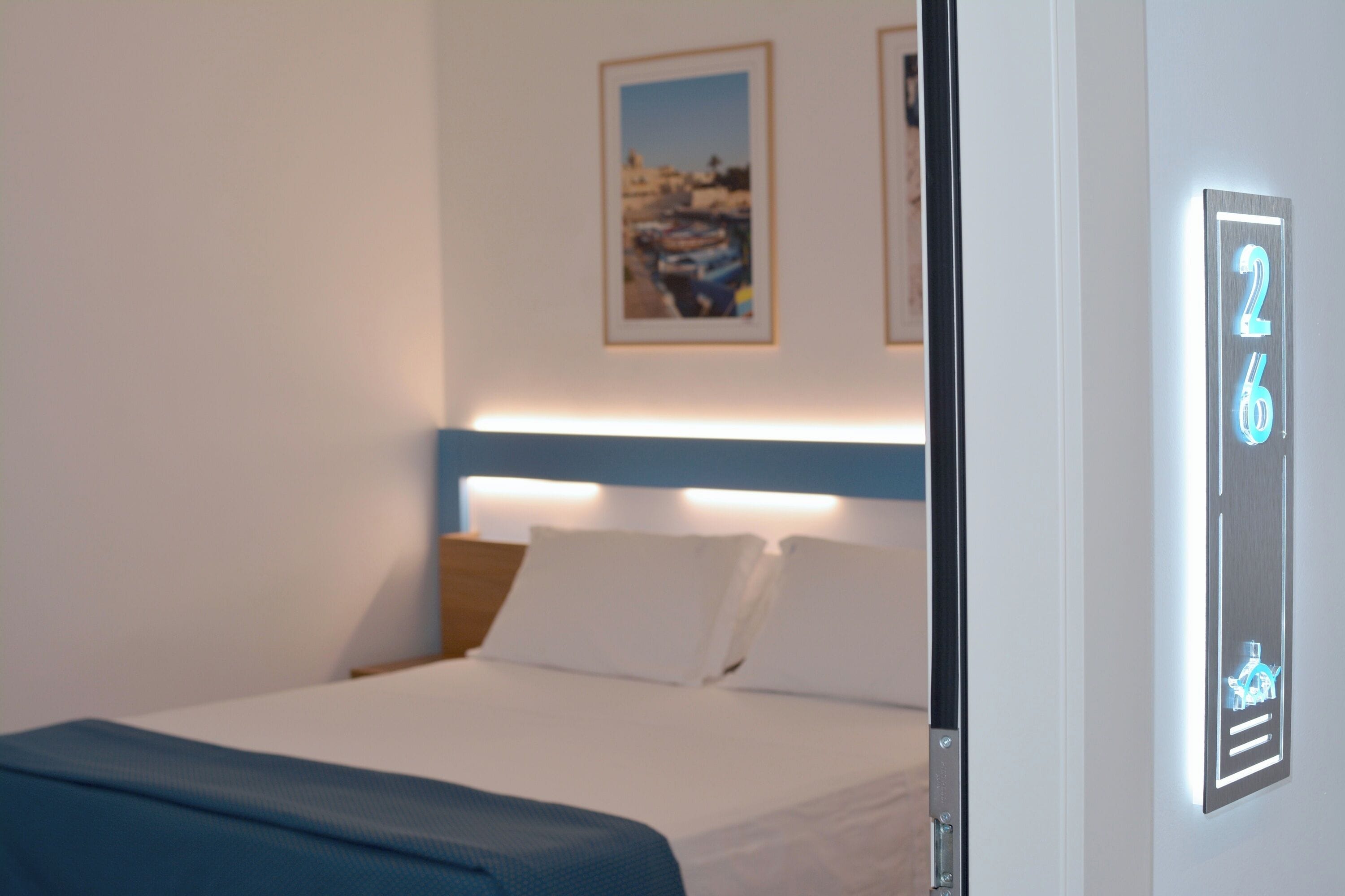 Hotellihuone, jossa on sänky, sängynpääty, seinällä oleva kehystetty kuva ja digitaalinen numeronäyttö.