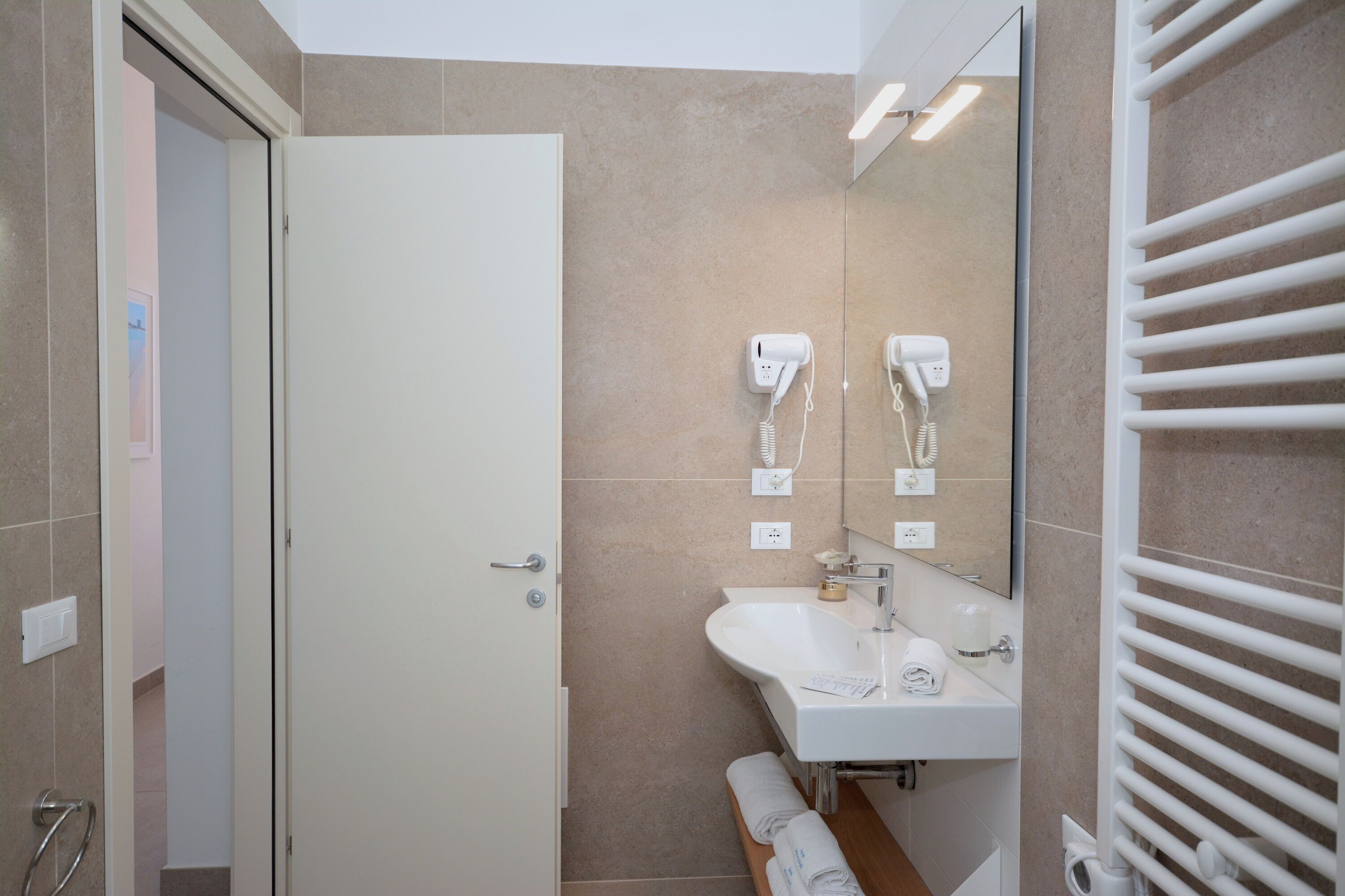 Un baño moderno con un lavamanos blanco, espejo y perchero. Hay dos secadores de pelo montados en la pared.