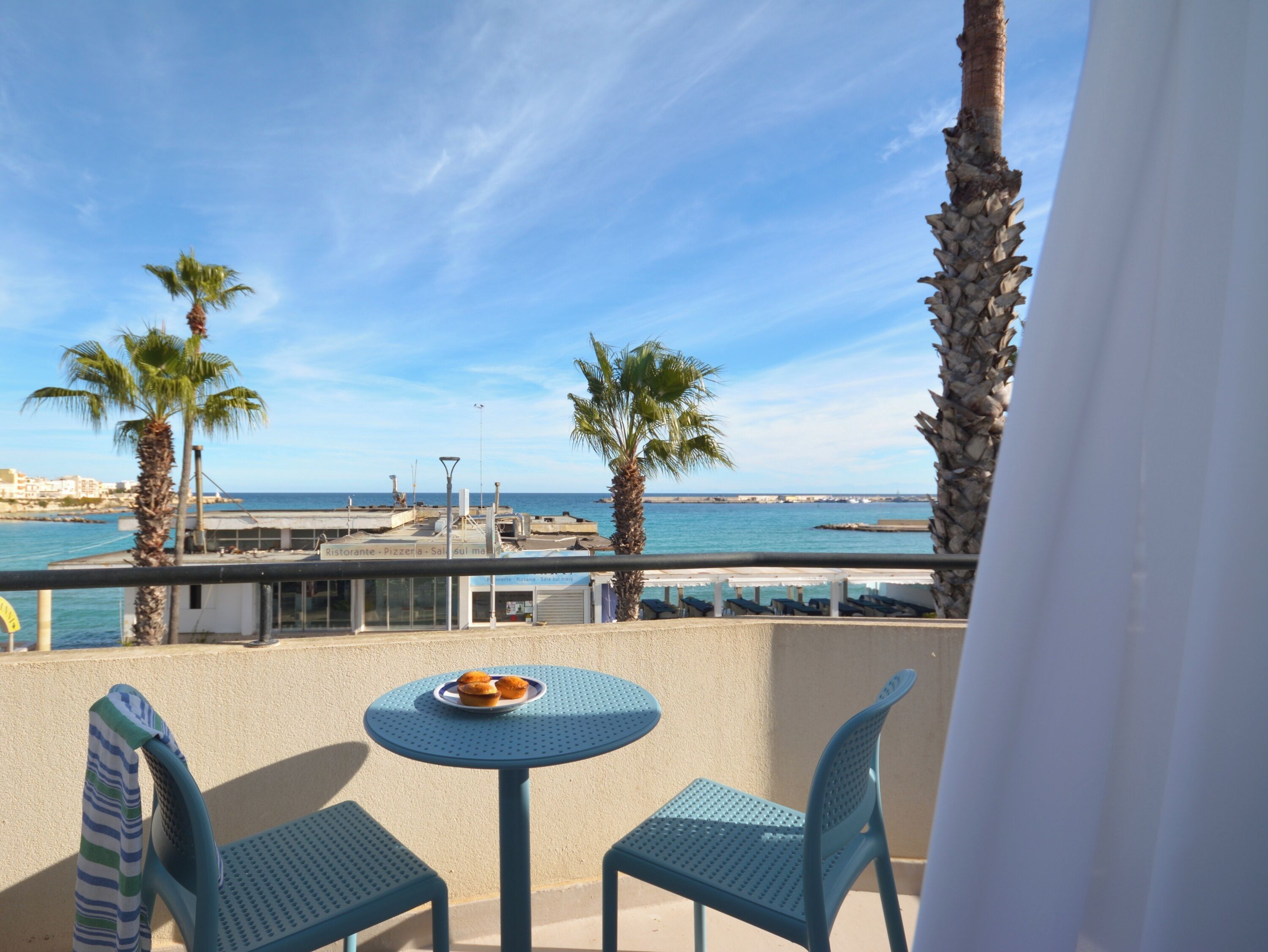 A balcony with two chairs and a small table, overlooking a coastal area with palm trees and a view of the sea.