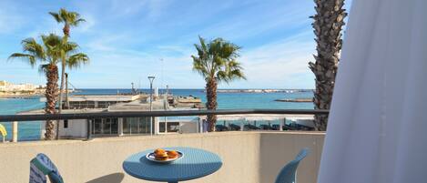A balcony with two chairs and a small table, overlooking a coastal area with palm trees and a view of the sea.