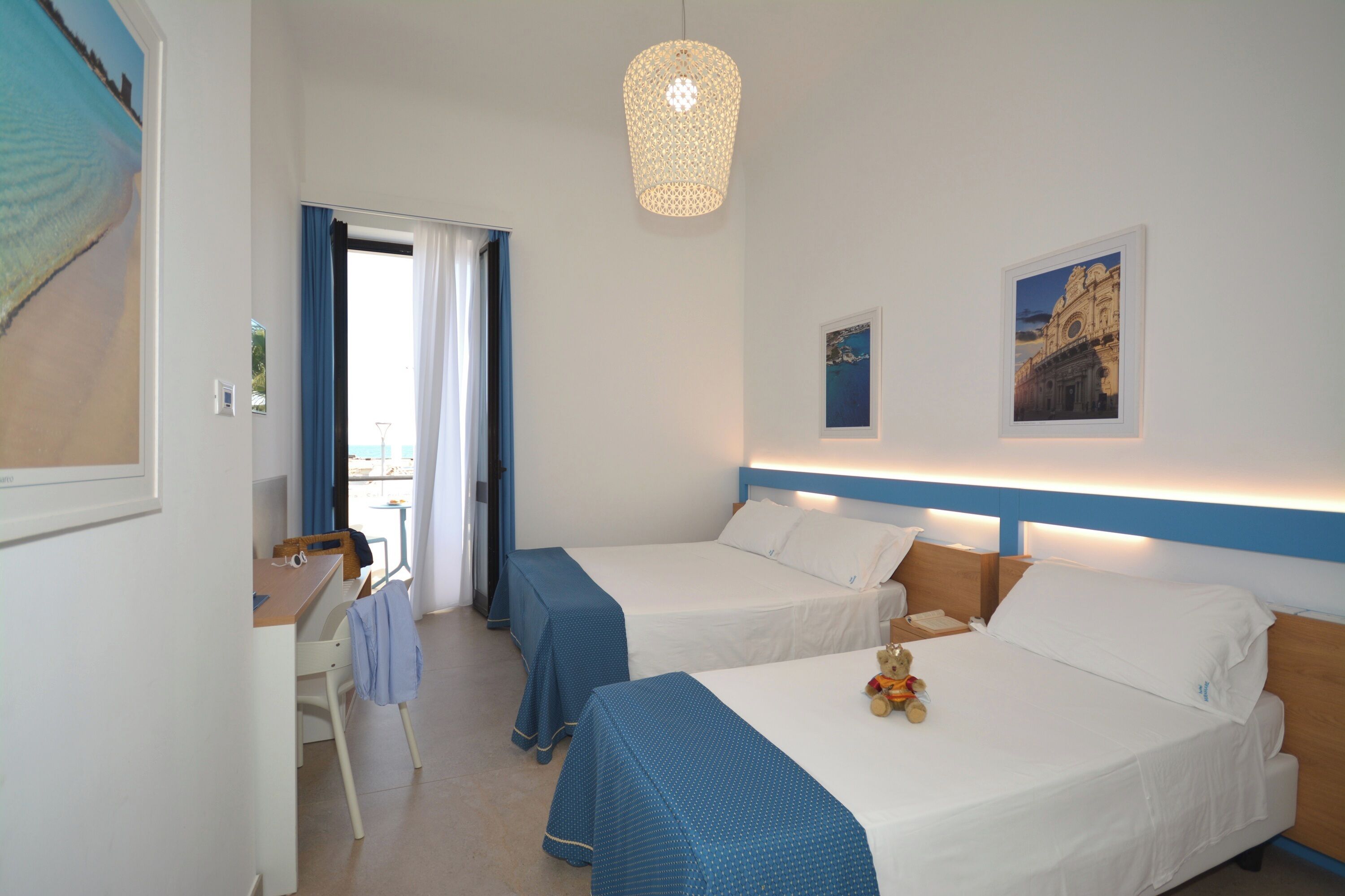 Habitación de hotel con dos camas, un escritorio y una silla. Tiene una ventana con cortinas, un aplique de pared y un cuadro en la pared.