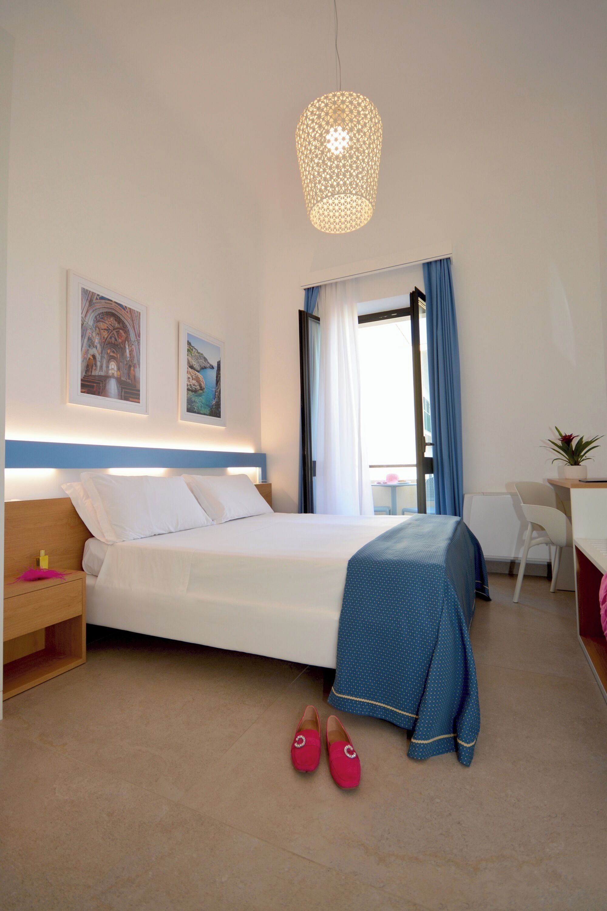 A hotel room with a bed, a desk, a chair, a small table with a plant, and a window with curtains.