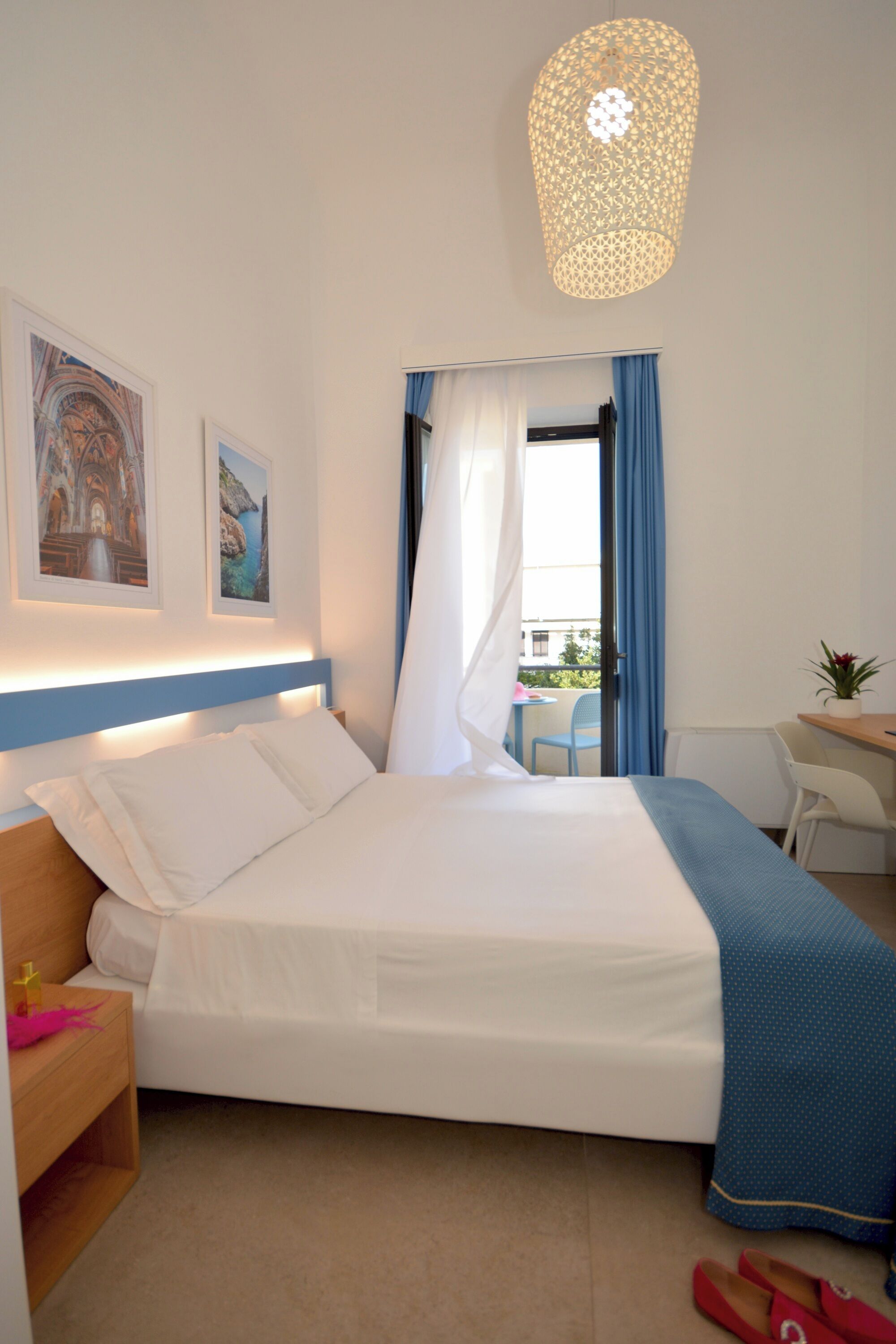 A hotel room with a bed, a desk, a chair, a window with curtains, and a hanging light fixture.
