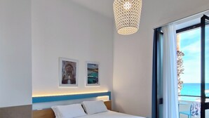 A hotel room with a bed, a nightstand, a desk with a hat and sunglasses, a hanging light fixture, and a balcony with a view of the sea. - Hotel Miramare (Otranto)