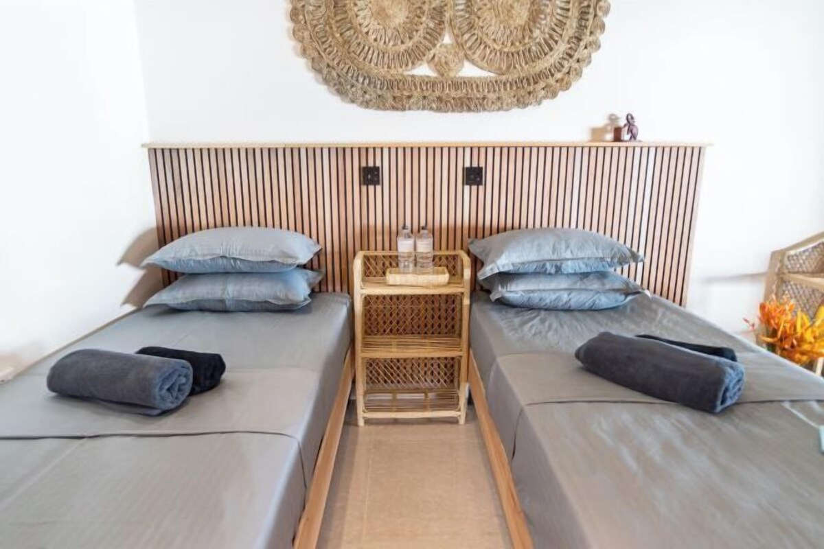 Une chambre avec deux lits, chacun doté d’un matelas et d’oreillers gris, d’une tête de lit en bois, d’une décoration murale tissée et d’une petite table en bois sur laquelle sont posées des bouteilles.