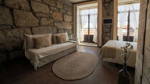 A cozy room with a stone wall, a sofa, a bed, and a large window with curtains. - Belos Aires Apartments (Porto)