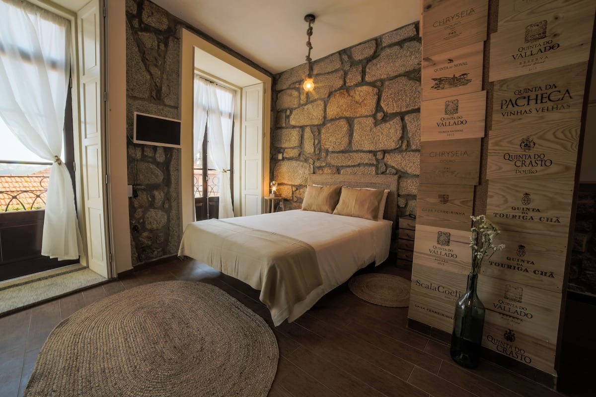 Un dormitorio con pared de piedra, cama con ropa de cama color beige, ventana grande con cortinas y televisor montado en la pared.