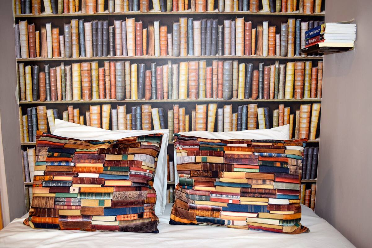 Un dormitorio con una cama, almohadas hechas de libros y una estantería llena de libros.