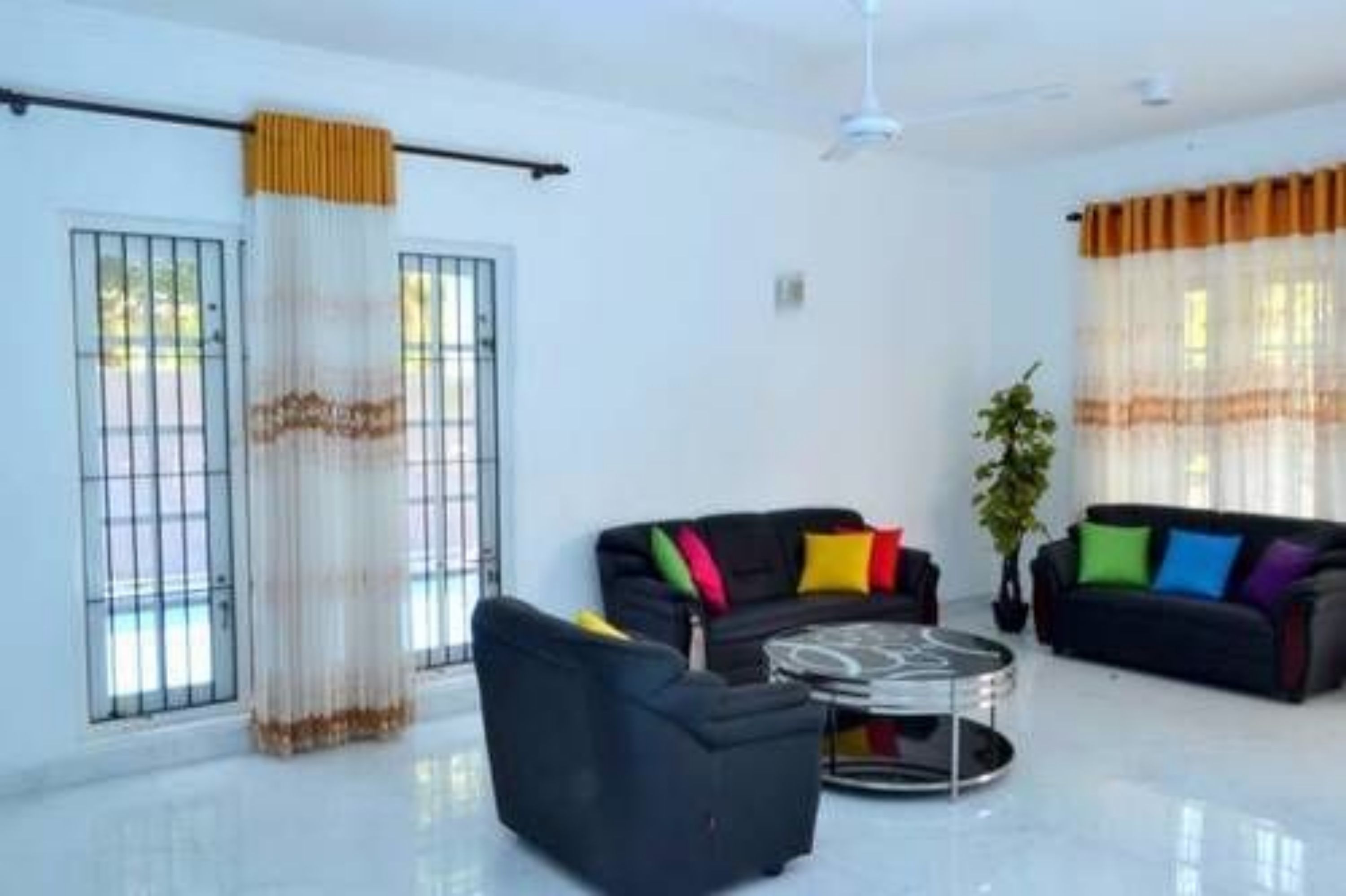A modern living room with a black sofa, colorful cushions, a glass coffee table, and sheer curtains.