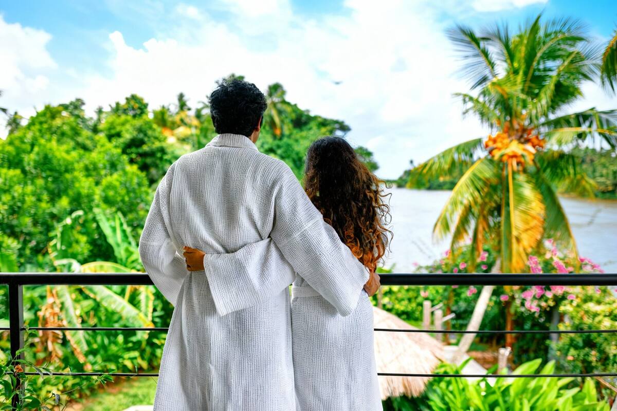 A couple standing on a balcony overlooking a lush garden with a river and palm trees.