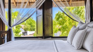 Una cama con sábanas y almohadas blancas, una puerta de madera y vistas a la vegetación a través de amplias ventanas.