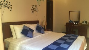 A neatly made bed with white and blue bedding, a wooden headboard, and a blue and white checkered blanket. There is a wooden dresser with a mirror and a lamp on the side. - Tra Que Riverside Homestay (Da Nang)