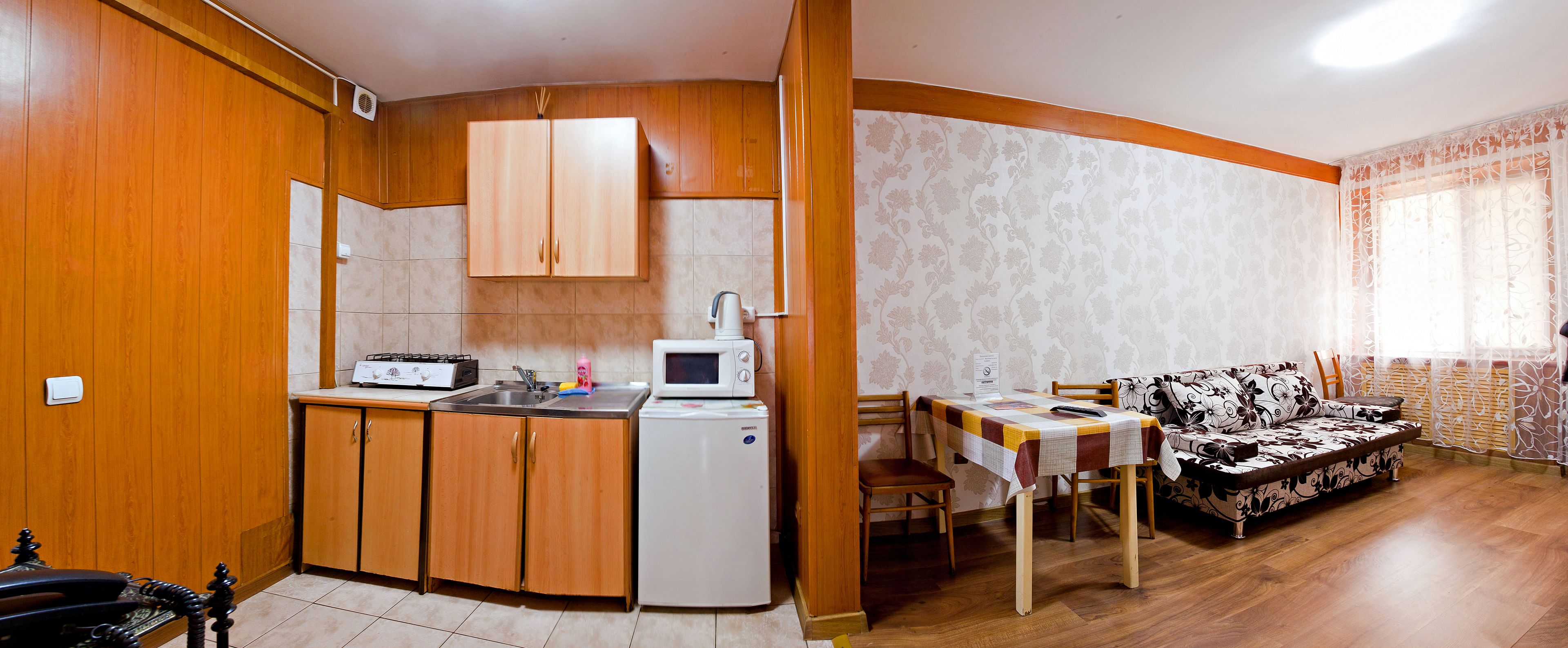 A compact kitchen with wooden cabinets, a microwave, and a small refrigerator. There is a dining area with a table and chairs, and a patterned sofa.