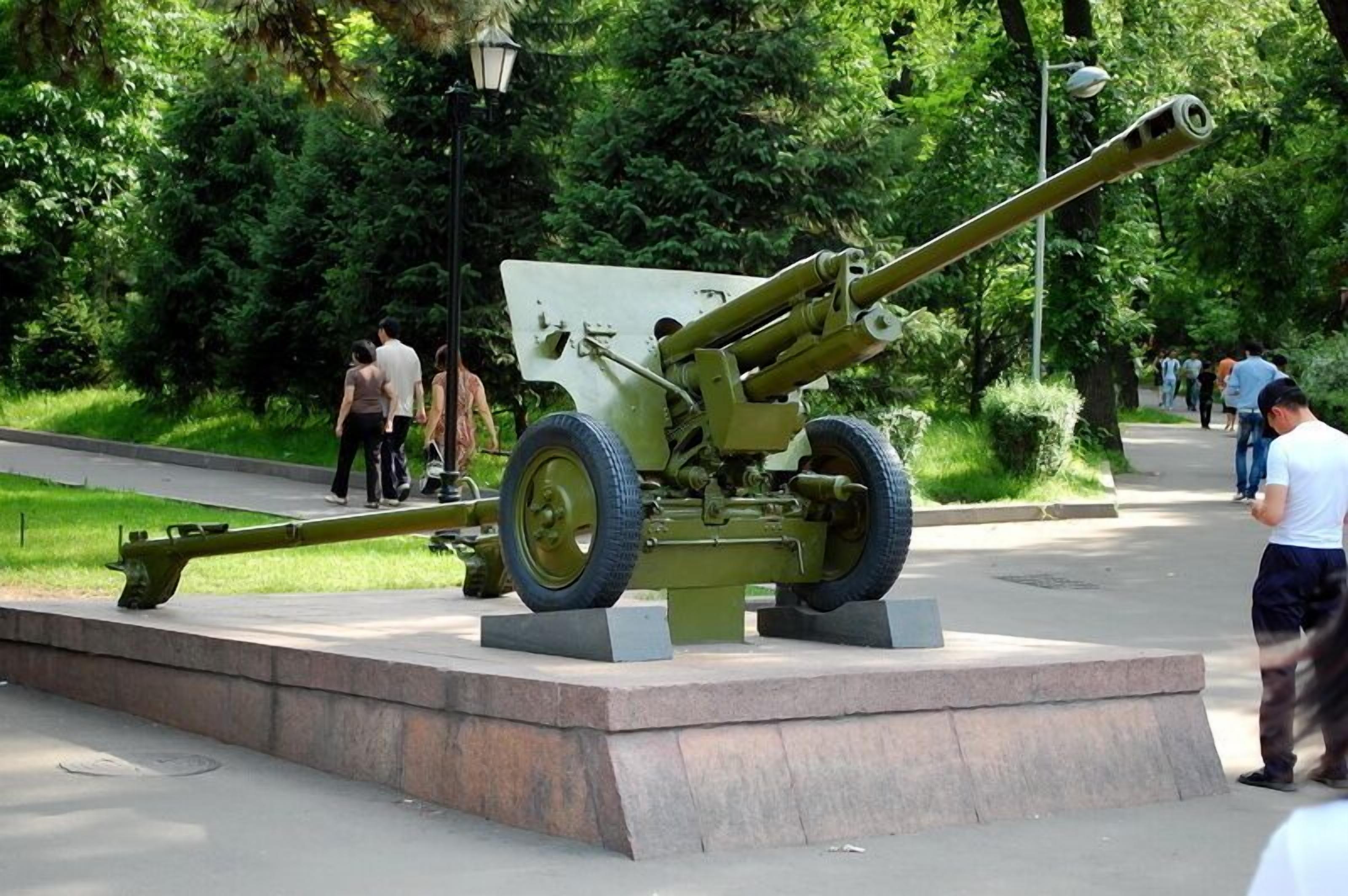 Un cañón militar exhibido en un parque con un sendero pavimentado y vegetación.