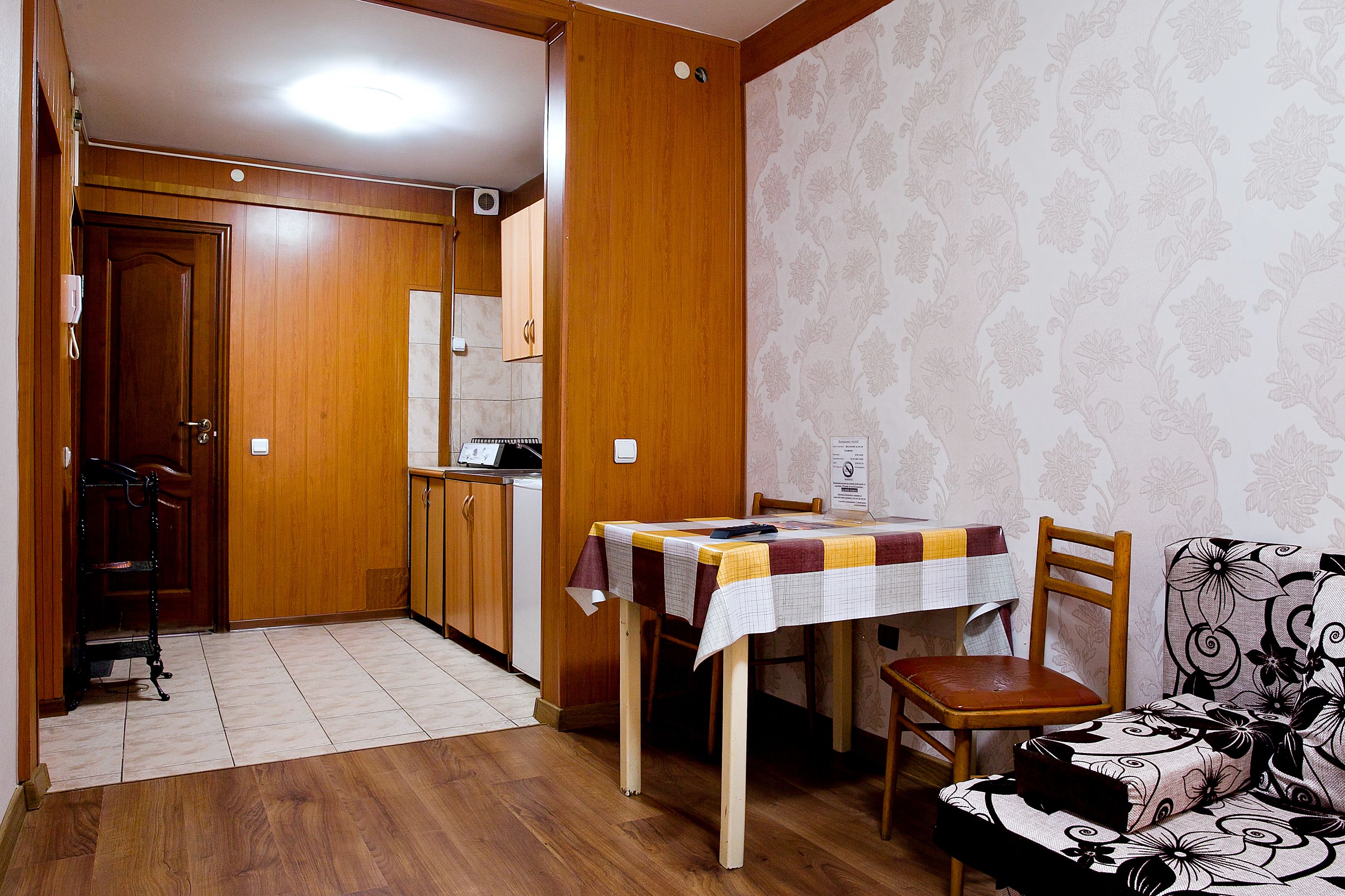A small, cozy room with a wooden floor, a dining table with a tablecloth, a sofa, and a kitchen area with wooden cabinets and a refrigerator.