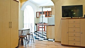 A kitchen with wooden cabinets, a black and white checkered floor, a dining area with stools, and a TV on a cabinet. - Lessor Apartments (Almaty)