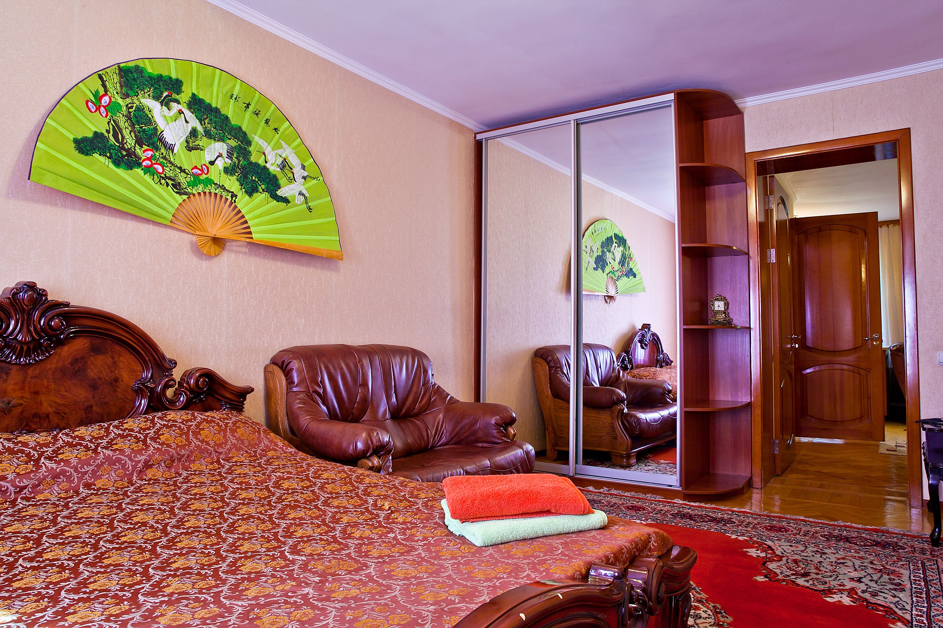 Une chambre à coucher avec un grand lit, un fauteuil en cuir, une armoire en bois et un ventilateur traditionnel fixé au mur.