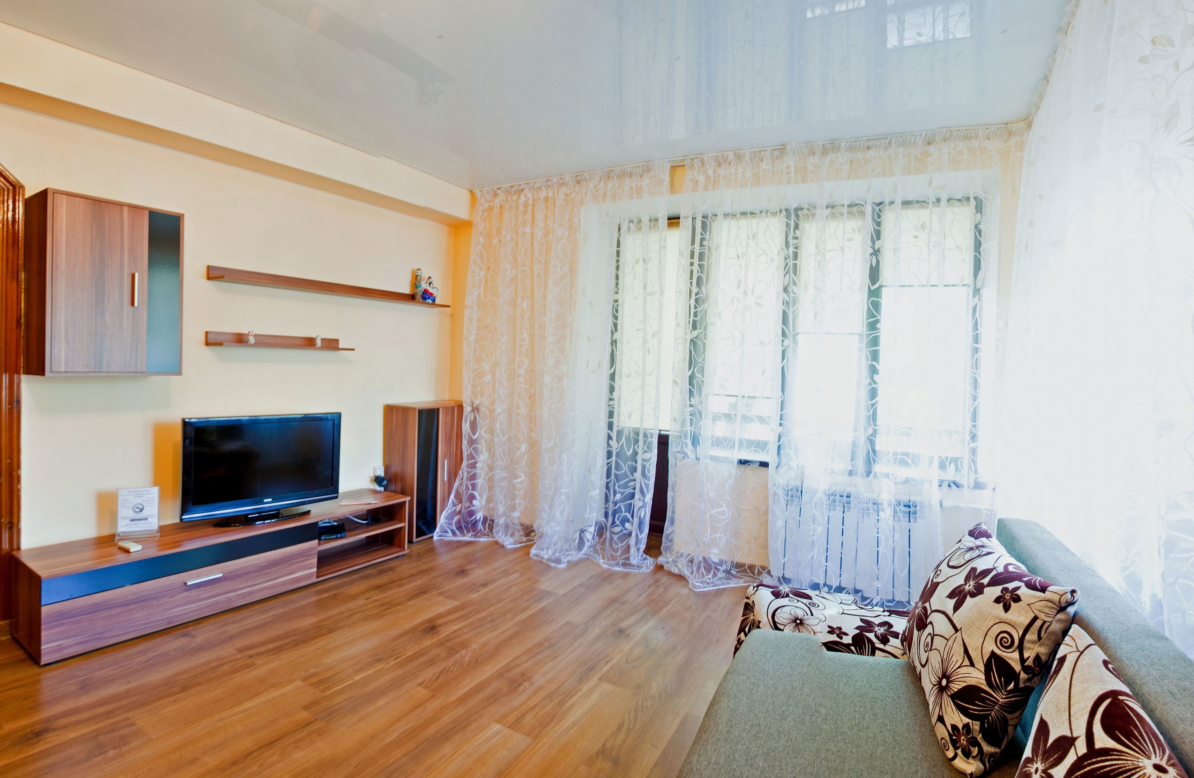 A living room with a flat-screen TV on a wooden stand, a sofa, and a window with sheer curtains.