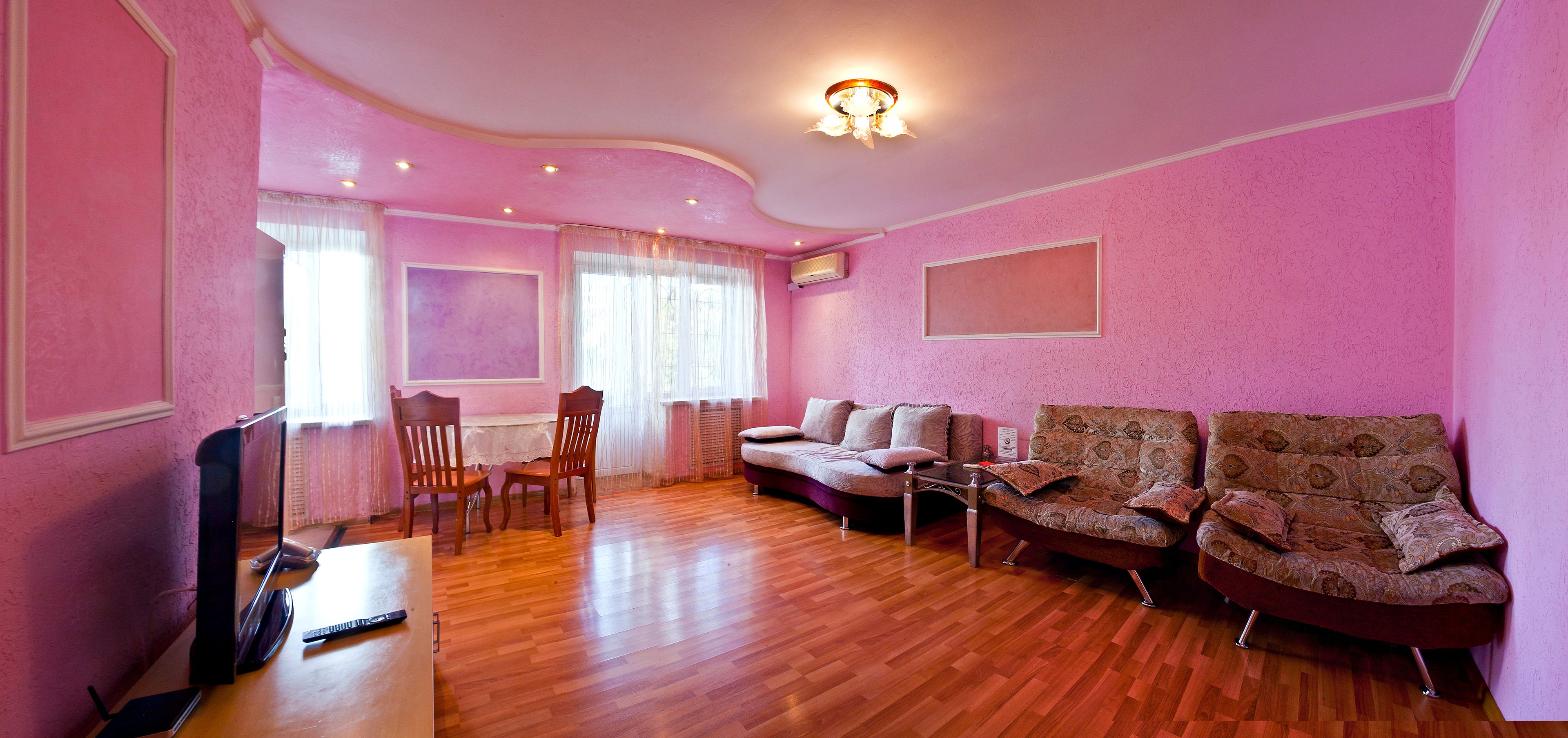 A pink-walled room with a wooden floor, a sofa, two armchairs, a dining table with chairs, and a TV.
