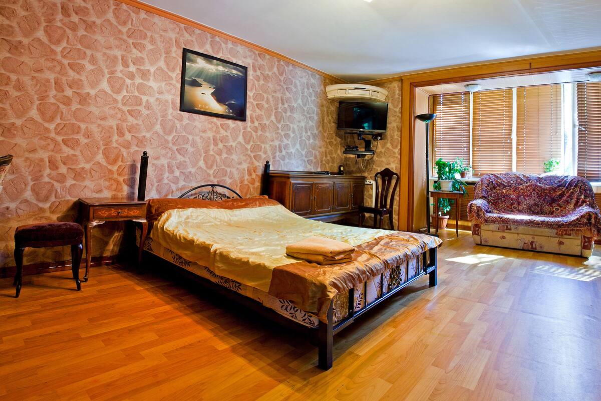A bedroom with a stone-patterned wall, a bed, a wooden dresser, a chair, and a television.