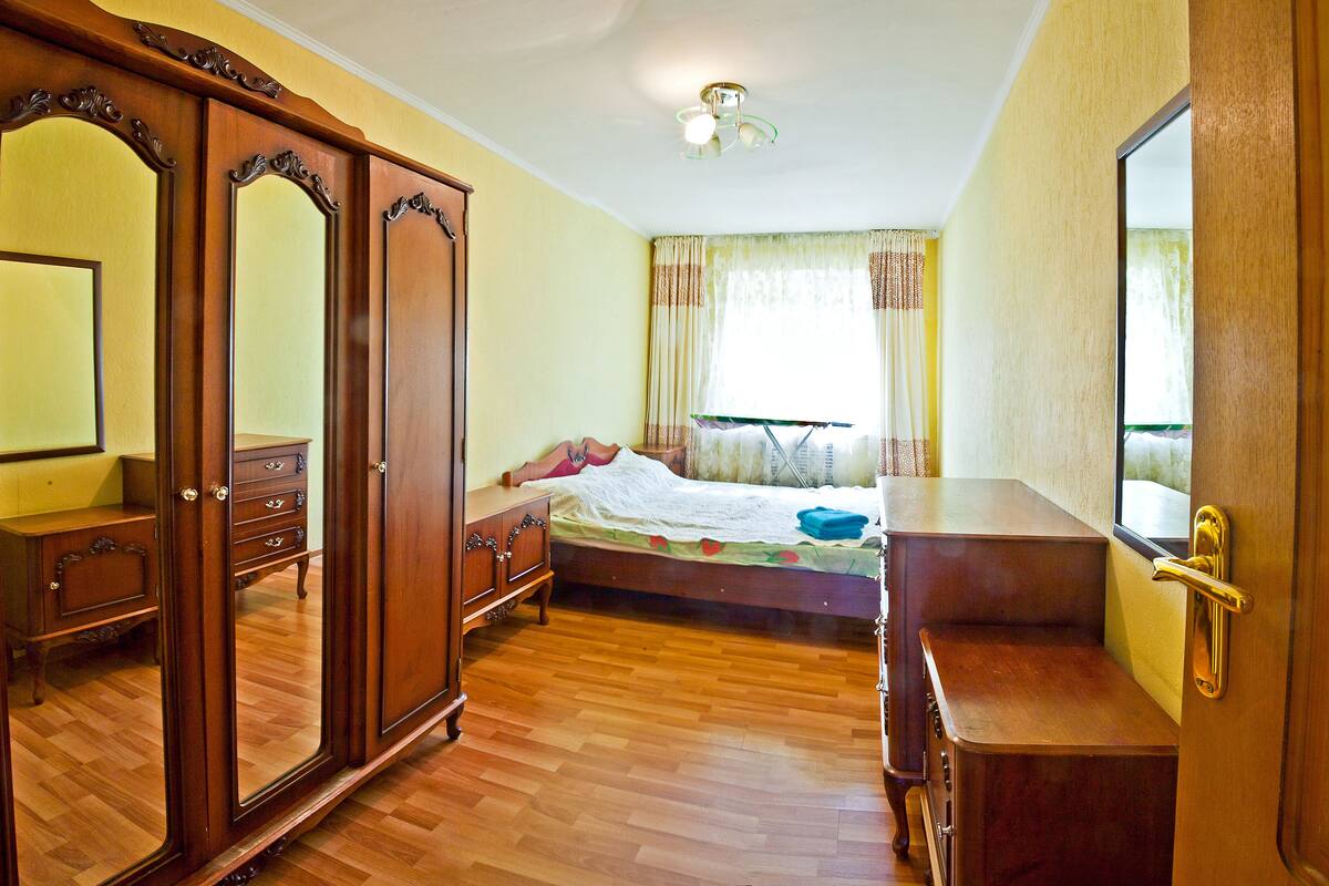 A bedroom with a wooden wardrobe, a bed with a white bedspread, and a window with curtains.