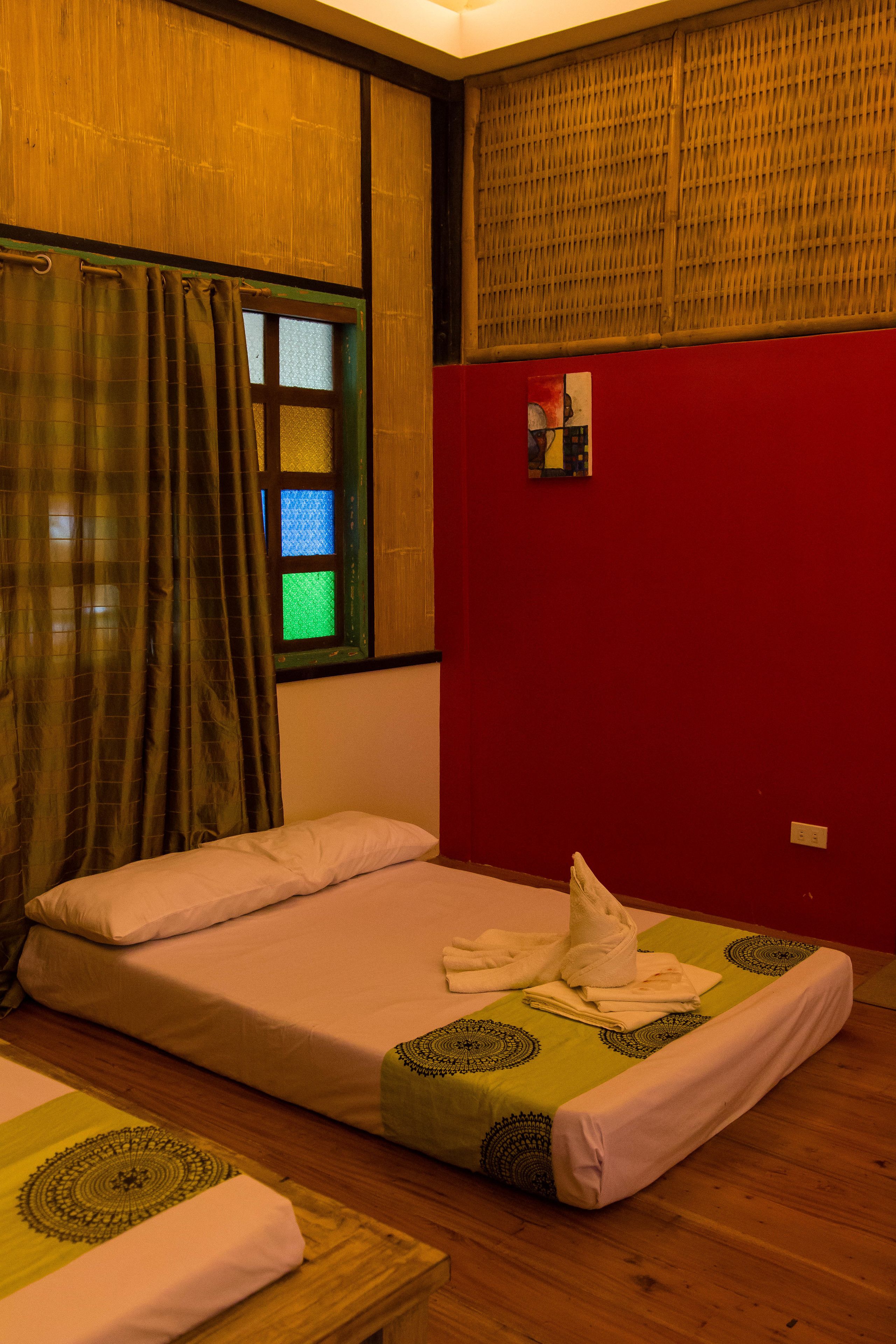 Une chambre avec un lit simple, une fenêtre avec un rideau, un mur rouge et un petit tableau.