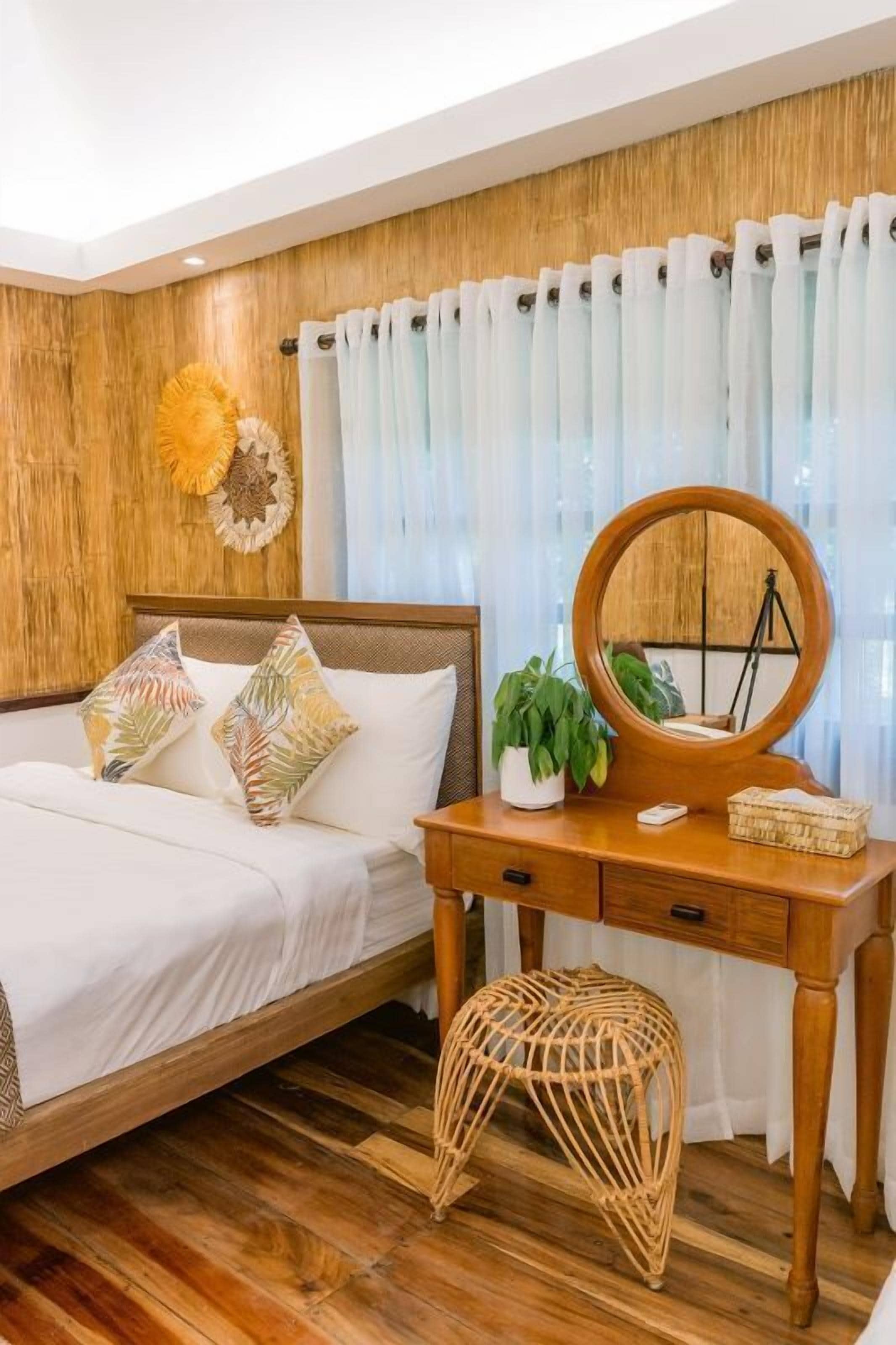Un dormitorio con cabecera de madera, cama con ropa de cama blanca, cómoda de madera con espejo y un taburete de mimbre.