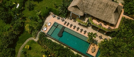 An aerial view of a resort with a swimming pool, lounge chairs, and a thatched-roof building.