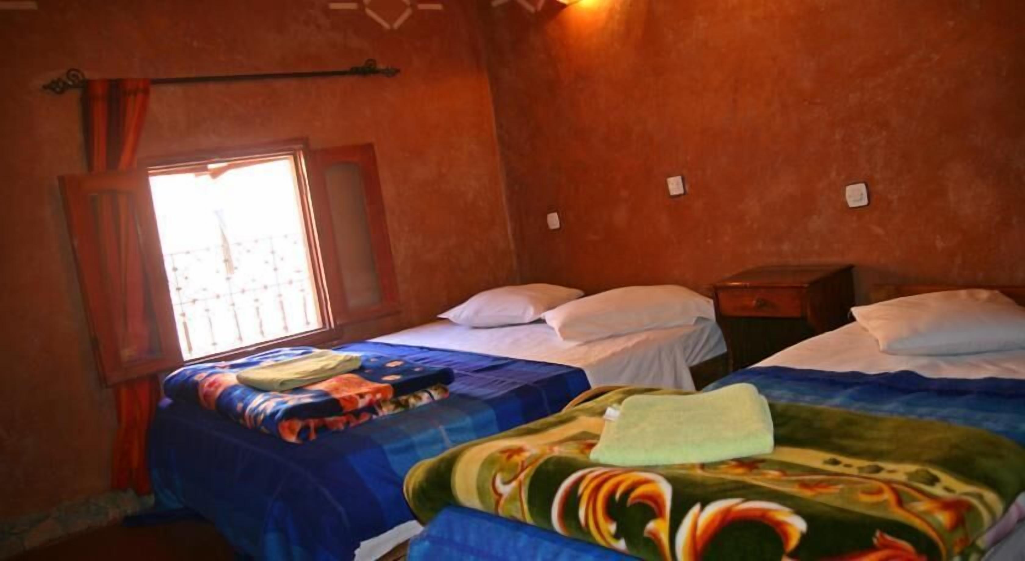 Ein Zimmer mit zwei Betten, jeweils mit einer blauen und bunten Decke. Es gibt ein Fenster mit einem Vorhang und einen kleinen Tisch mit einer Lampe.