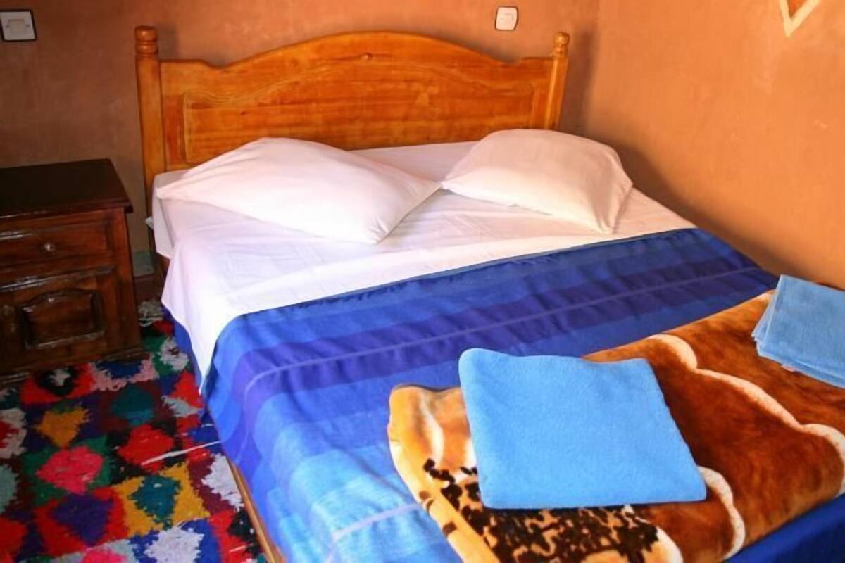 Cama com cabeceira de madeira, almofadas brancas, manta azul e duas toalhas dobradas.